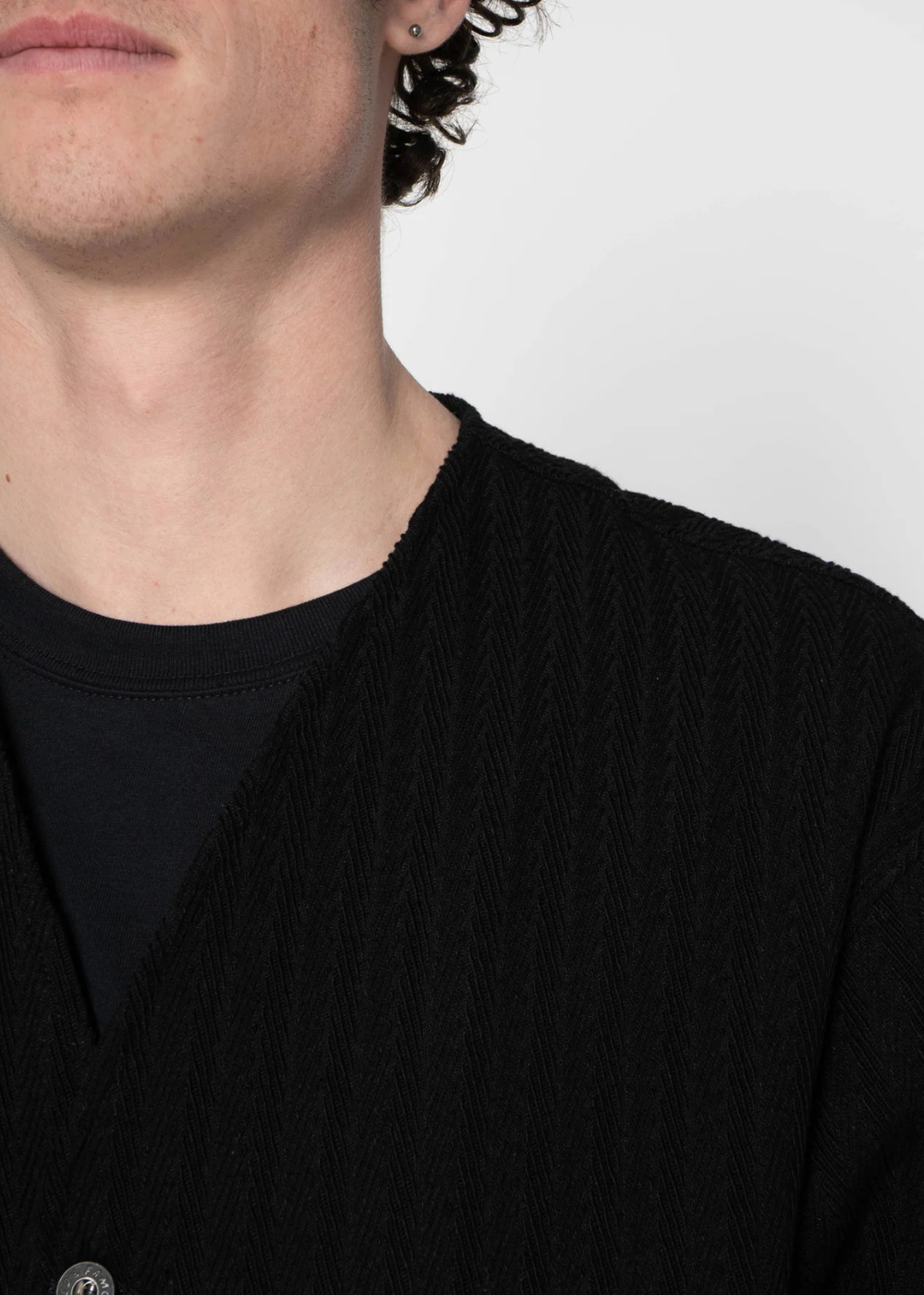 Close-up of a person wearing a black textured knit cardigan over a black crewneck t-shirt against a plain light gray background.