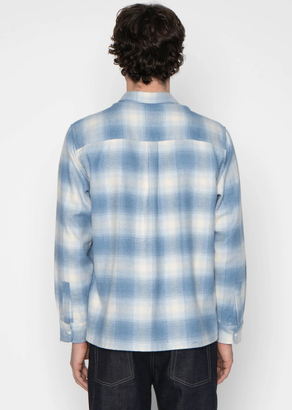 A man viewed from behind wearing a light blue and white plaid long-sleeve button-up shirt and dark jeans, against a plain white studio background.