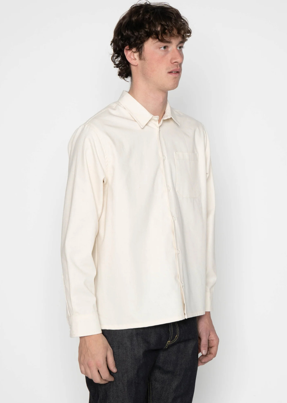 A man wearing a cream-colored long-sleeve button-down shirt and dark jeans, standing against a plain white studio background.