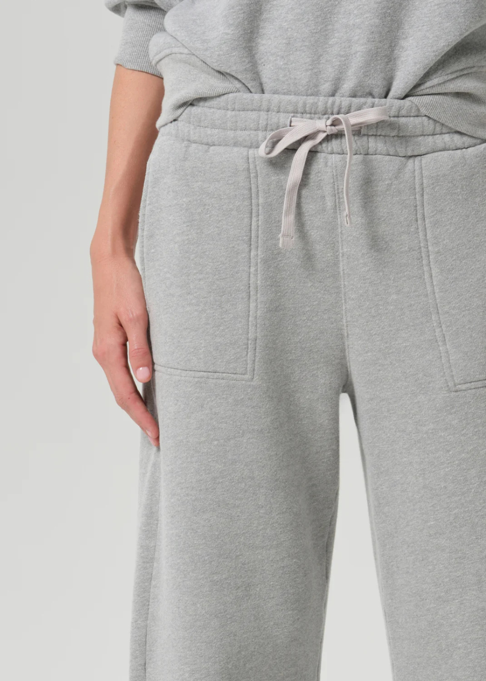 Close-up of a person wearing light gray heather sweatpants with a drawstring waist and front patch pockets, paired with a matching gray sweatshirt.