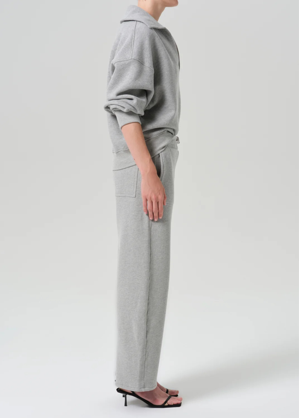 A model shown from the side wearing a light gray ribbed knit hoodie and matching wide-leg trousers, paired with black heeled sandals, against a light gray studio background.
