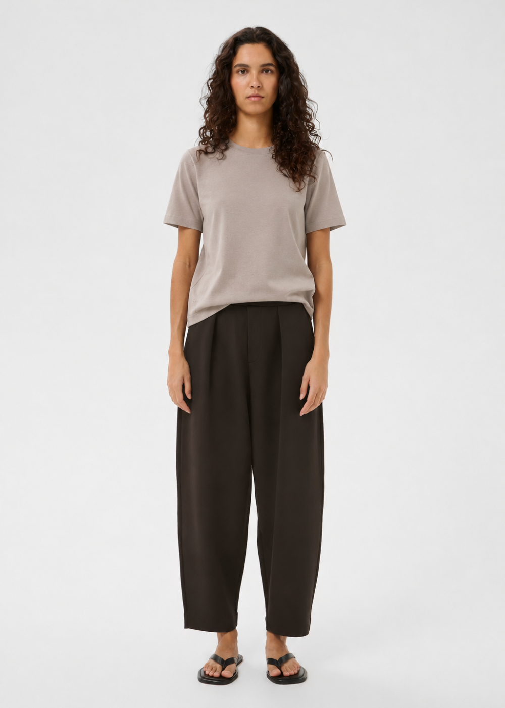 A woman stands against a light gray studio background wearing a plain taupe short-sleeve T-shirt, dark charcoal wide-leg cropped pants, and black sandals.
