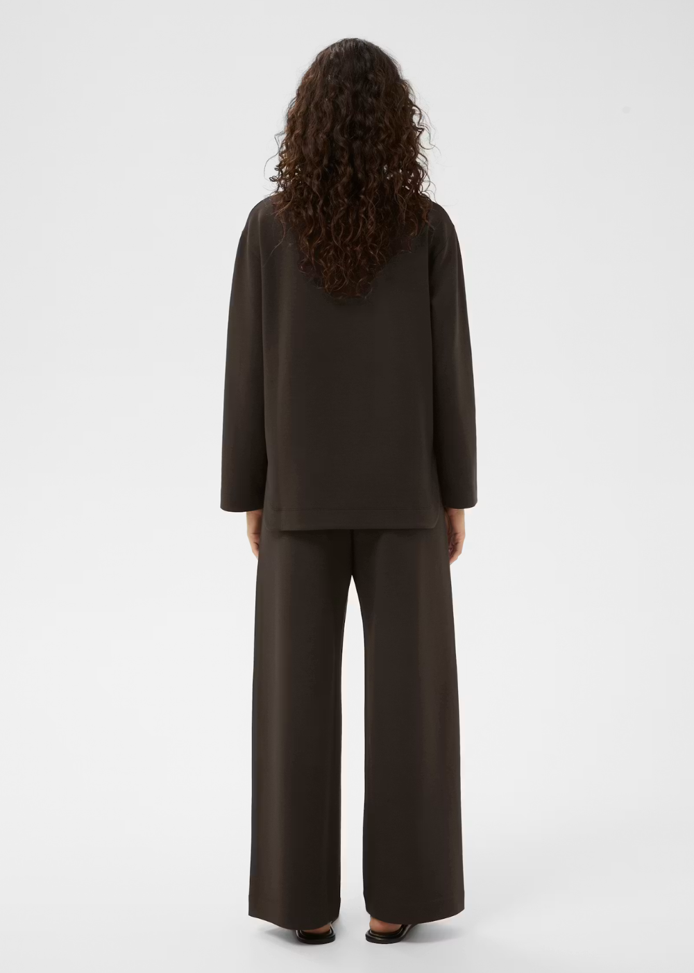 A model viewed from the back wearing a dark brown long-sleeve top and matching wide-leg trousers against a plain light gray studio background.