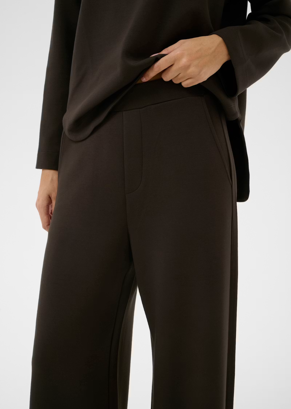 A close-up of a model wearing dark brown wide-leg trousers and a matching dark brown top, with one hand pulling the hem of the top.