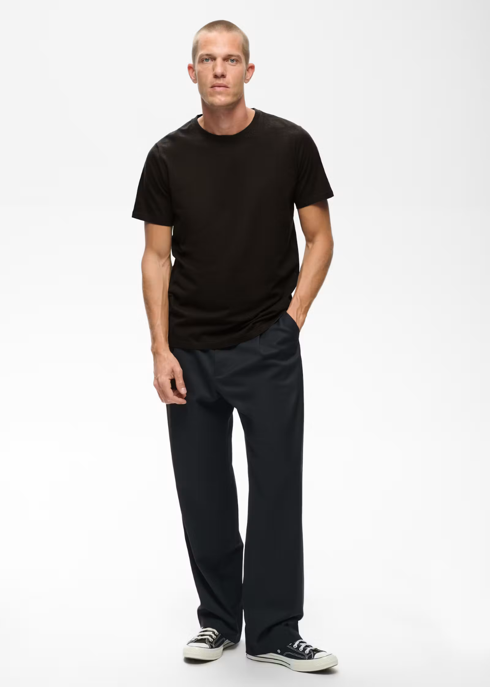A man modeling a black short-sleeve crewneck t-shirt and dark charcoal gray pants, standing against a plain white studio background.