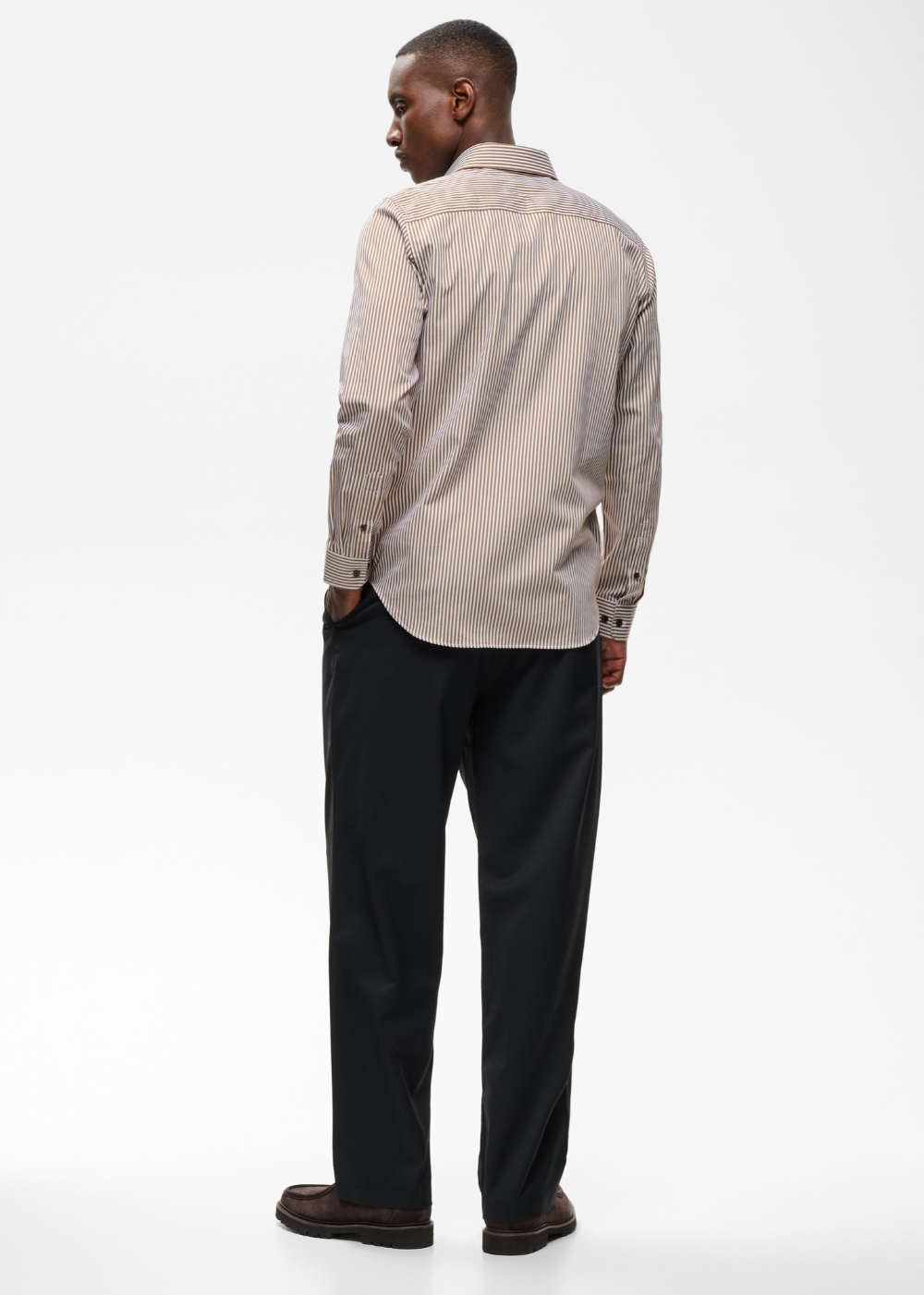 A man shown from the back wearing a light beige and white vertically striped long-sleeve button-down shirt, dark trousers, and brown shoes, photographed against a plain white studio background.