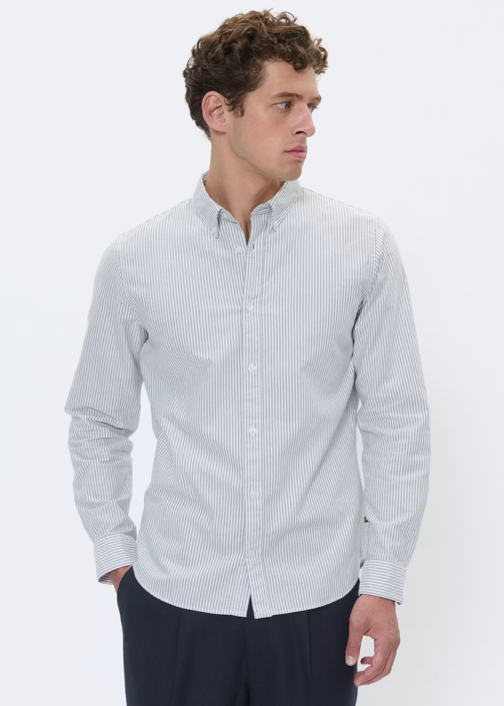A man wearing a light blue and white vertically striped long-sleeve button-down shirt, paired with dark trousers, photographed against a plain white studio background.