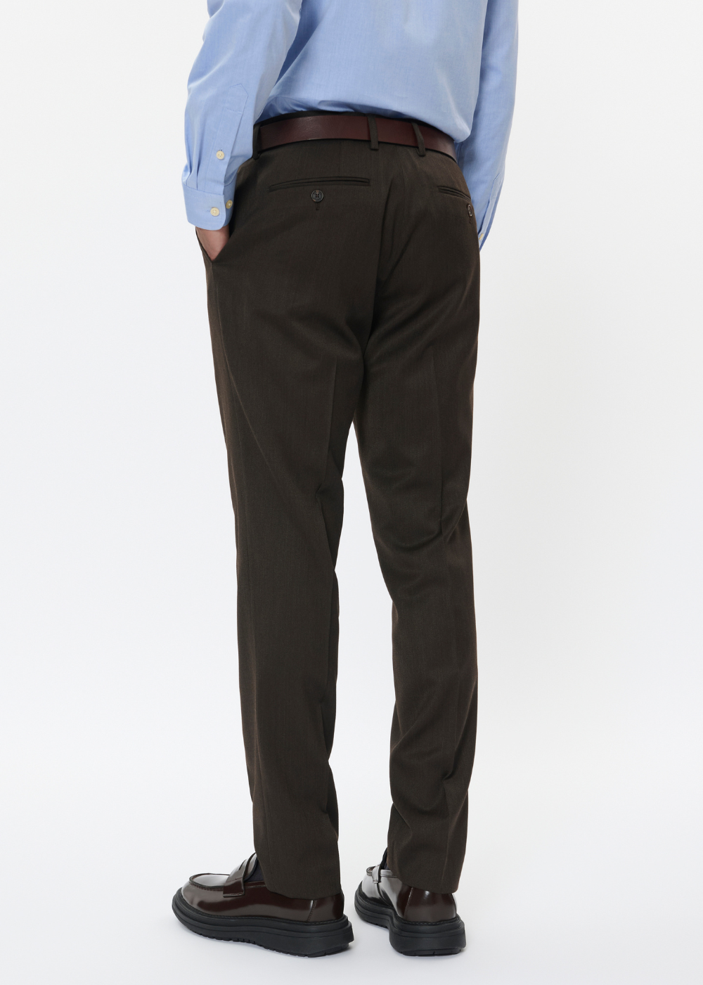 Rear view of a person wearing dark brown dress pants with a brown belt, a light blue dress shirt, and dark brown dress shoes, against a plain white studio background.