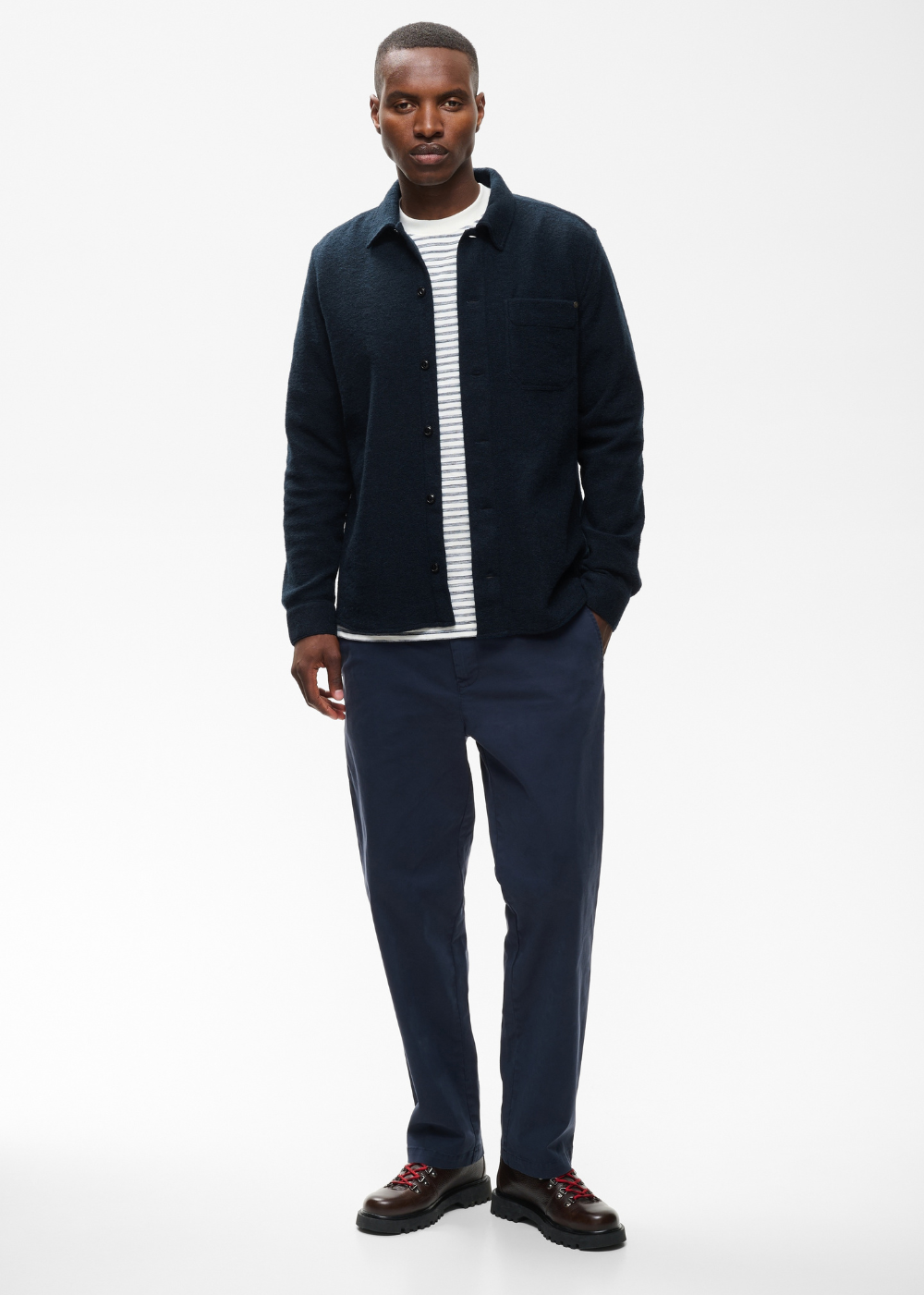 A man modeling a dark navy blue overshirt over a white and navy striped t-shirt, paired with navy trousers and dark loafers, against a plain white studio background.