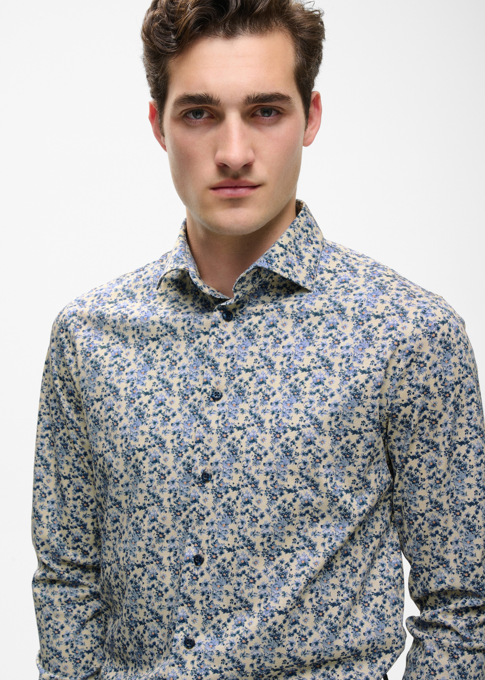 A man wearing a long-sleeve button-down shirt with a dense blue and black floral print on a light beige background, photographed against a plain light gray studio background.