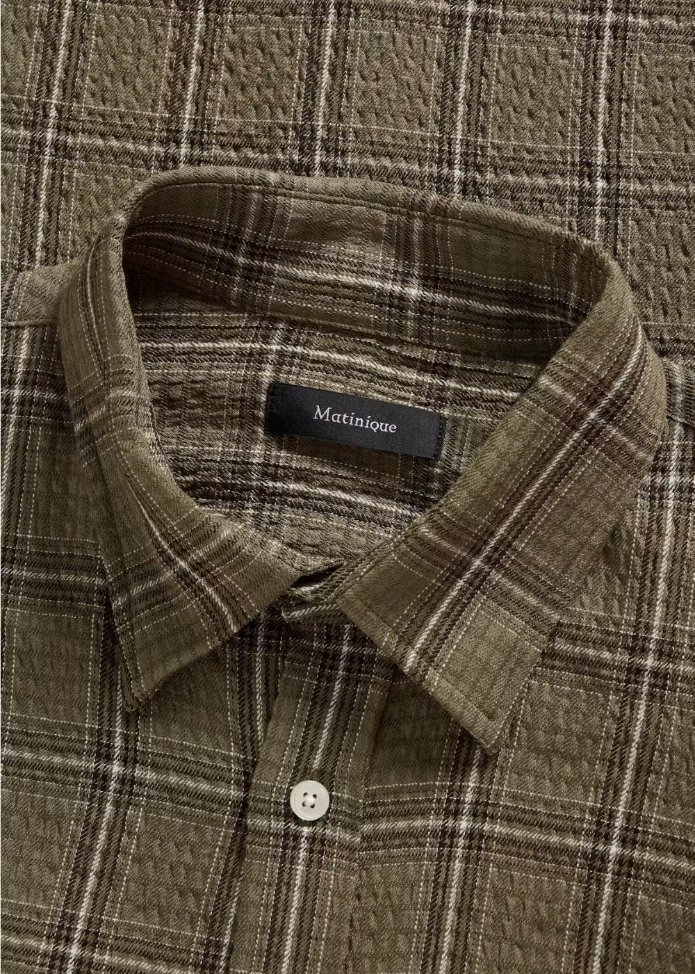 A close-up of a collared button-up shirt with a textured waffle-weave fabric in an olive green and brown plaid pattern. A black neck label reads 'Matinique'.