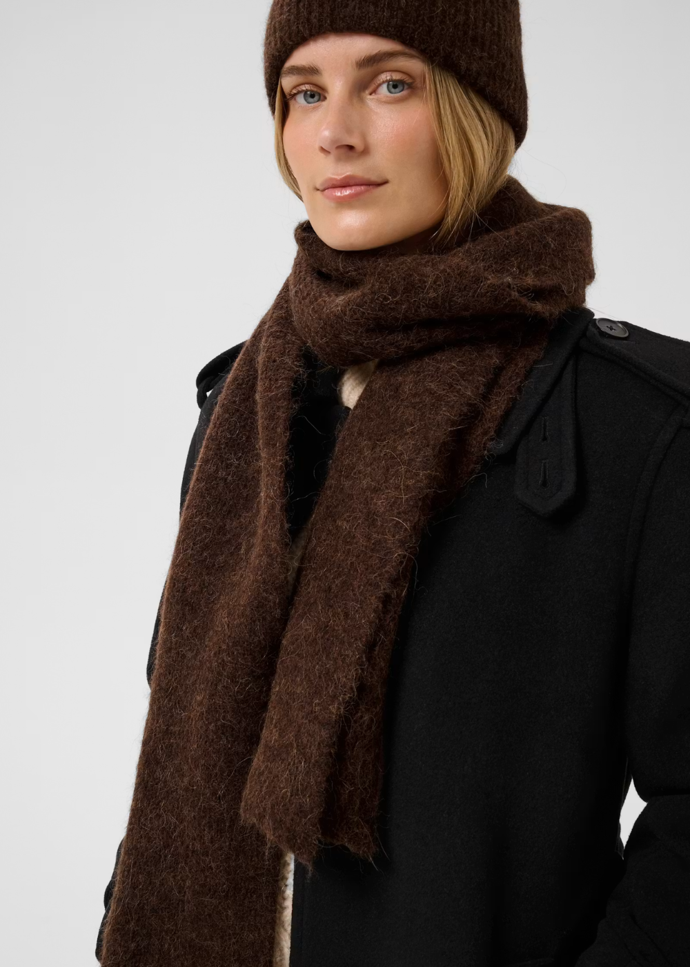 A woman wearing a thick, fuzzy dark brown scarf and a matching brown knit beanie, paired with a black coat, photographed against a light gray studio background.