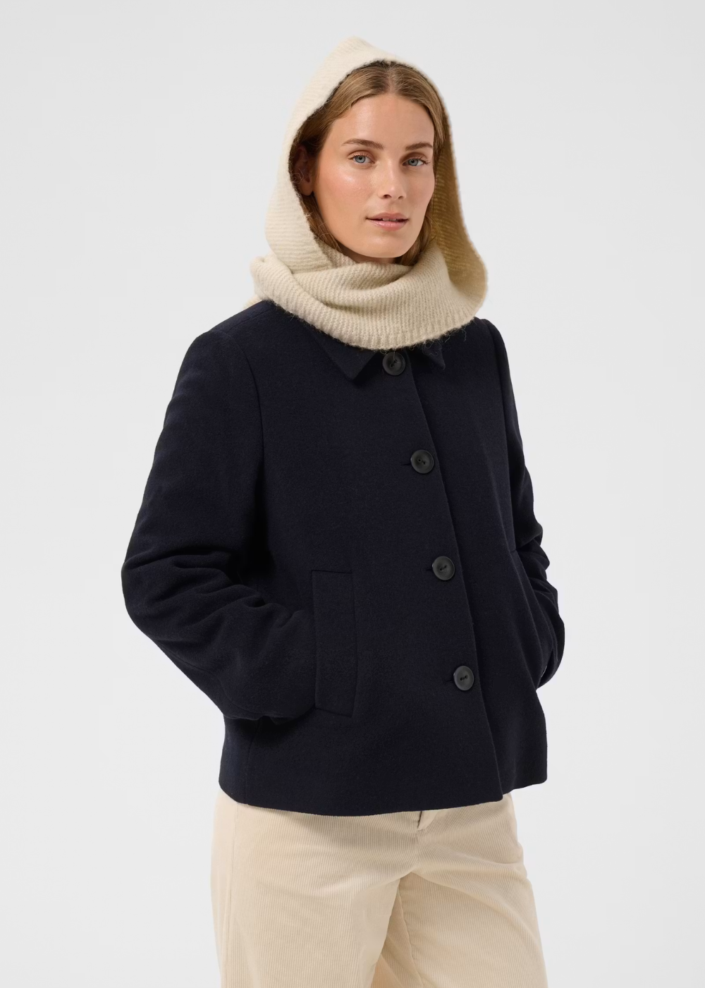 A woman wearing a dark navy wool-blend coat with a collar and front buttons, paired with a cream knit hood and matching cream pants, photographed against a light gray studio background.