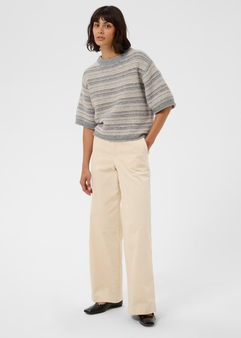A model stands against a light gray studio background wearing a loose, short-sleeve striped knit top in gray and beige tones, paired with wide-leg cream pants and black flats.