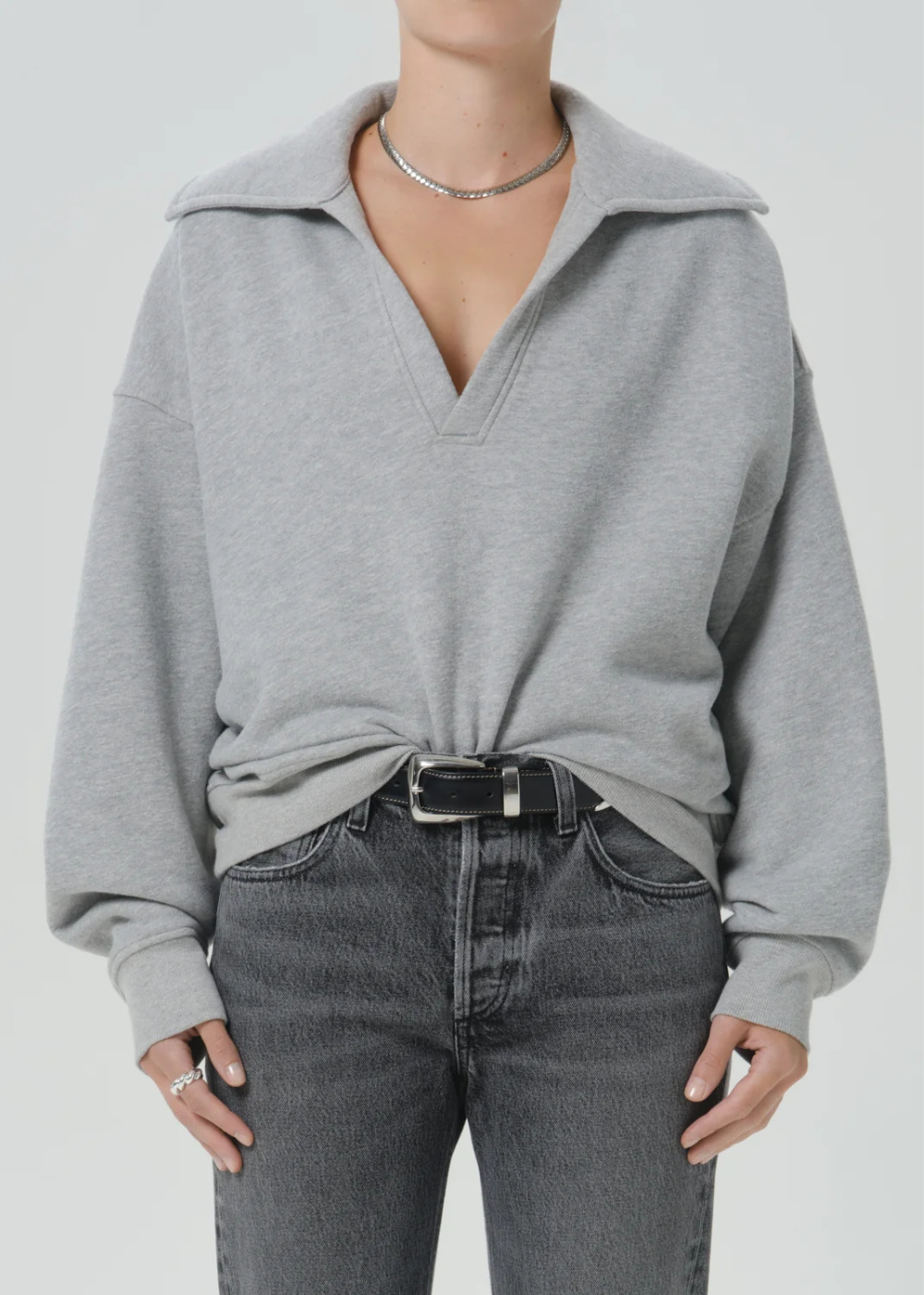 A person wearing an oversized light gray knit quarter-zip pullover with a wide collar, tucked into dark gray jeans with a black belt, against a plain light gray studio background.
