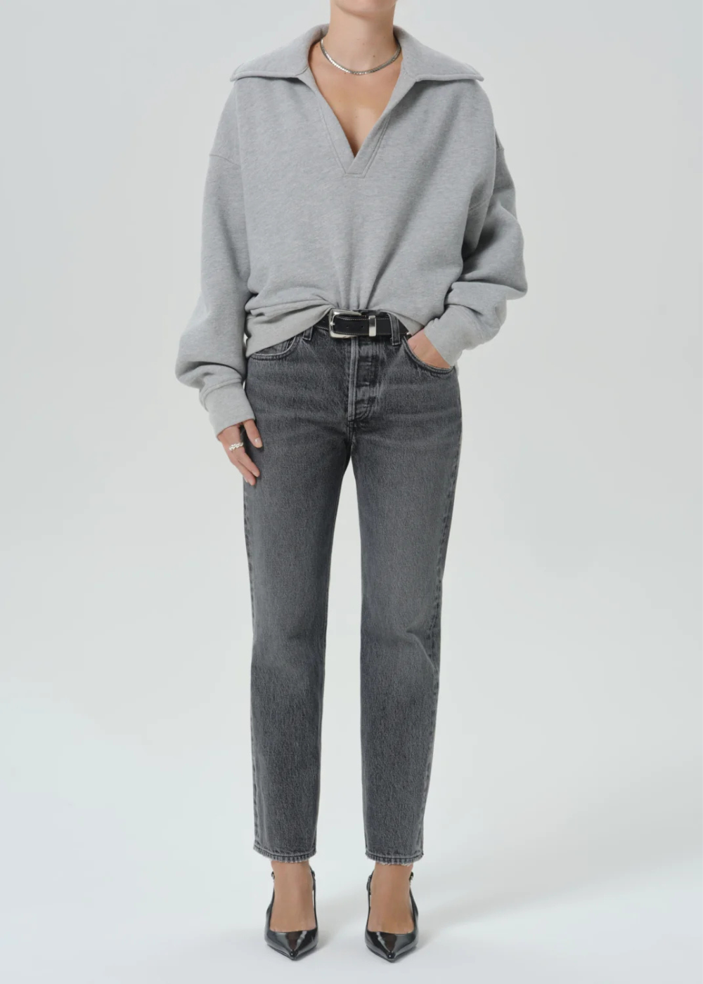 A model wearing a light gray oversized knit sweater with a collar, tucked into dark gray straight-leg jeans with a black belt, paired with black pointed-toe heels, against a light gray studio background.