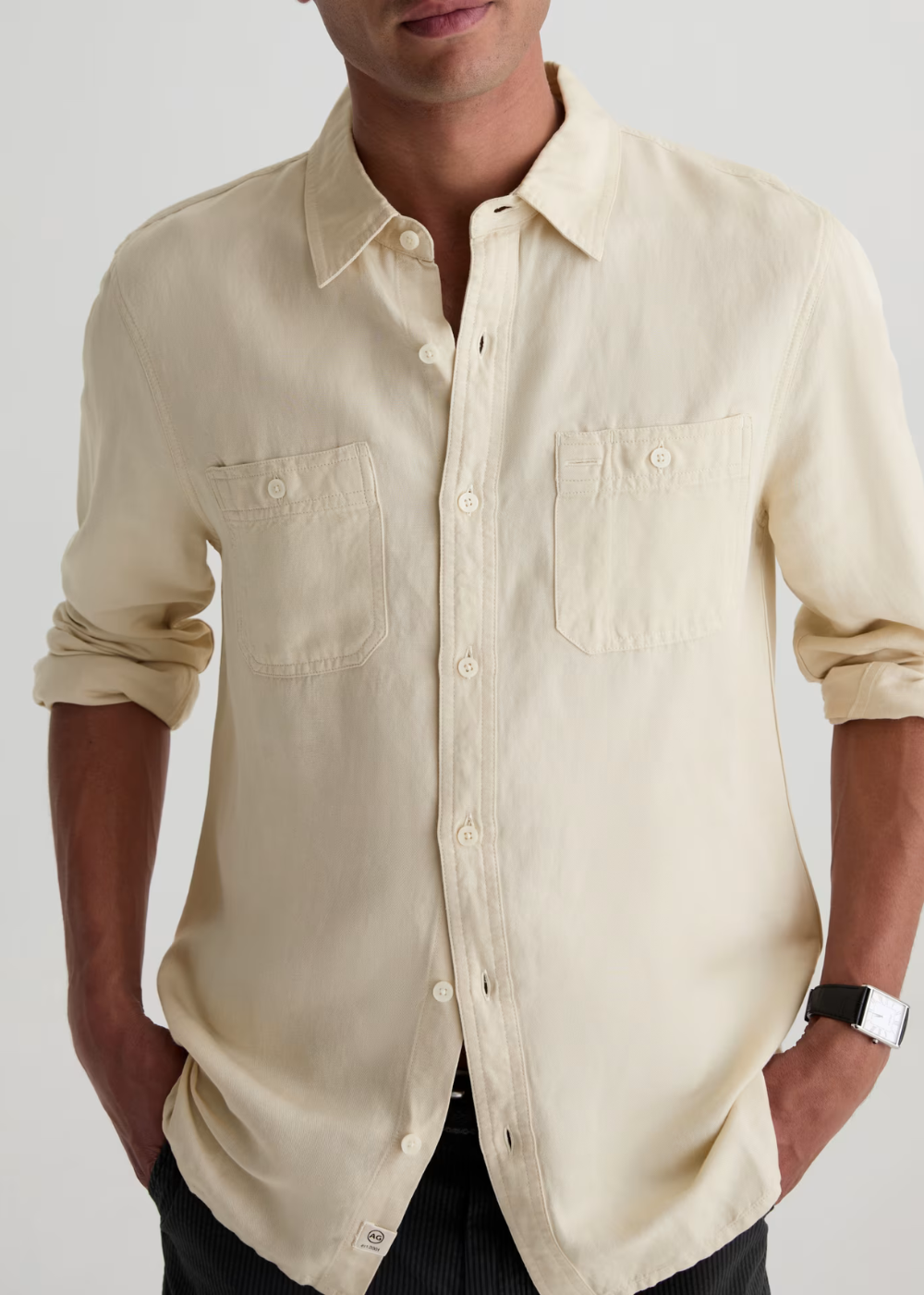 A man wearing a light cream-colored long-sleeve linen button-down shirt with two chest pockets, shown from the neck down against a light gray background.