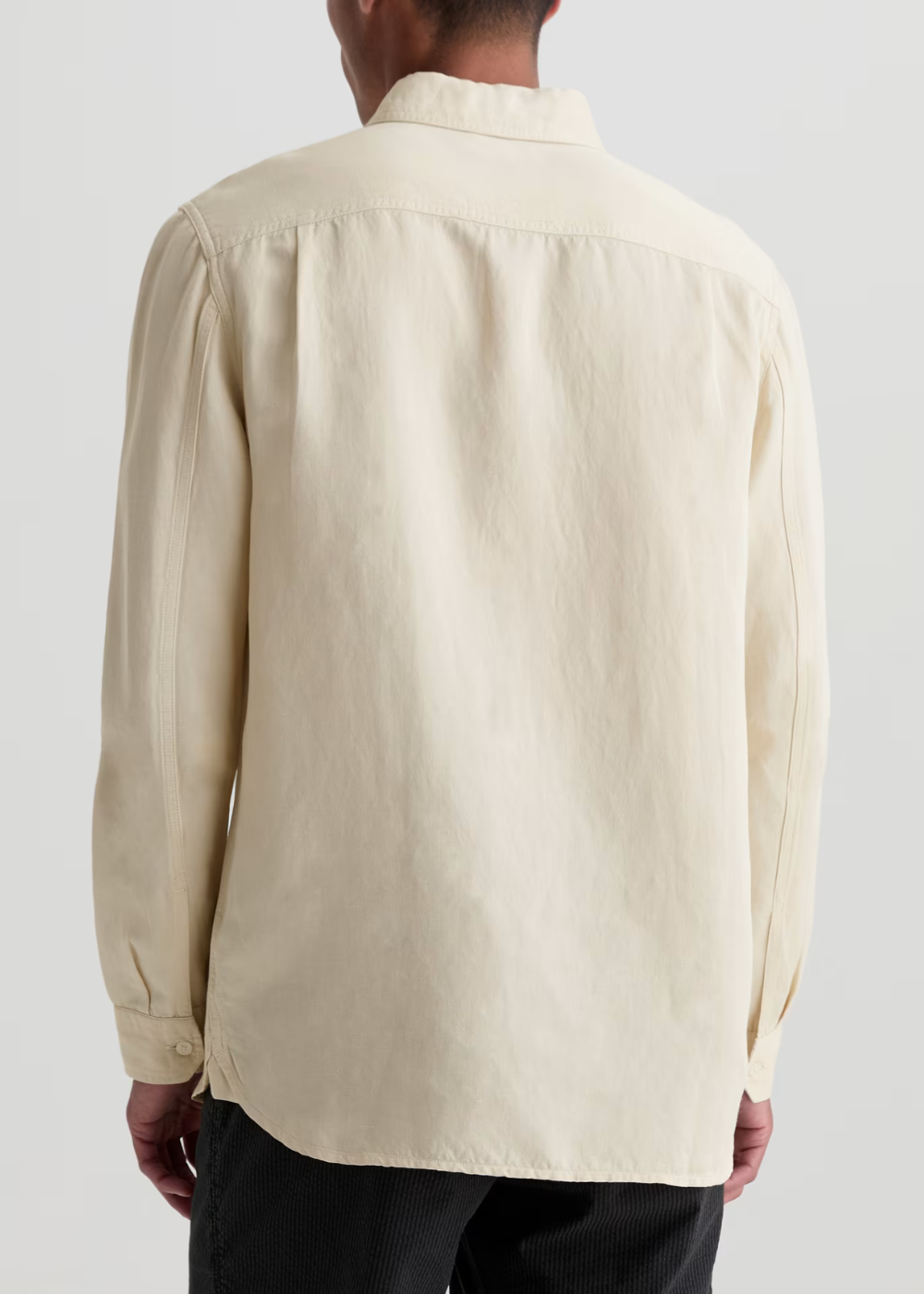 A man viewed from the back wearing a light cream-colored long-sleeve button-down shirt and dark trousers, set against a plain light gray studio background.