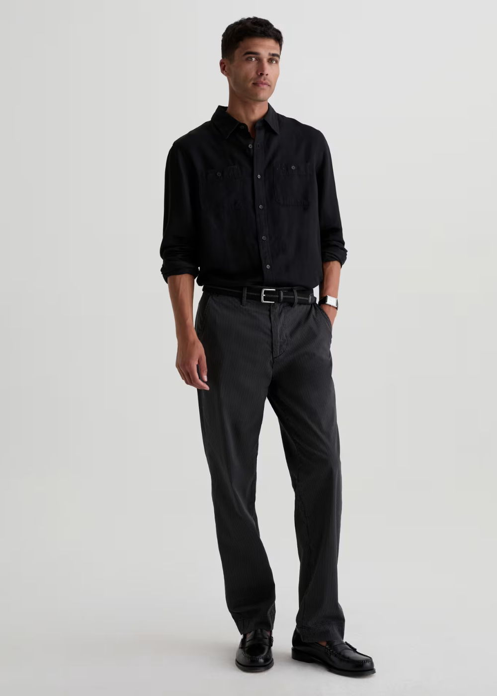 A man modeling a black long-sleeve button-down shirt and dark charcoal gray trousers, paired with a black belt and black leather loafers, against a plain light gray studio background.