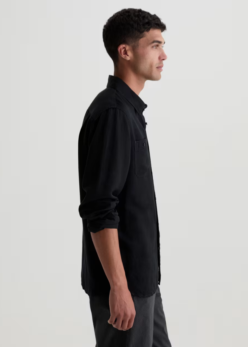 A man in side profile wearing a black long-sleeve button-up shirt and dark gray trousers, photographed against a plain light gray studio background.