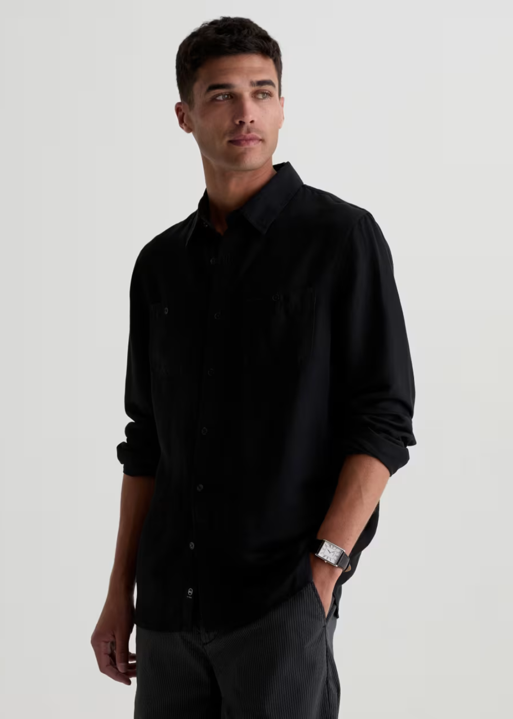 A man wearing a black long-sleeve button-up shirt and dark trousers, standing against a plain light gray studio background.