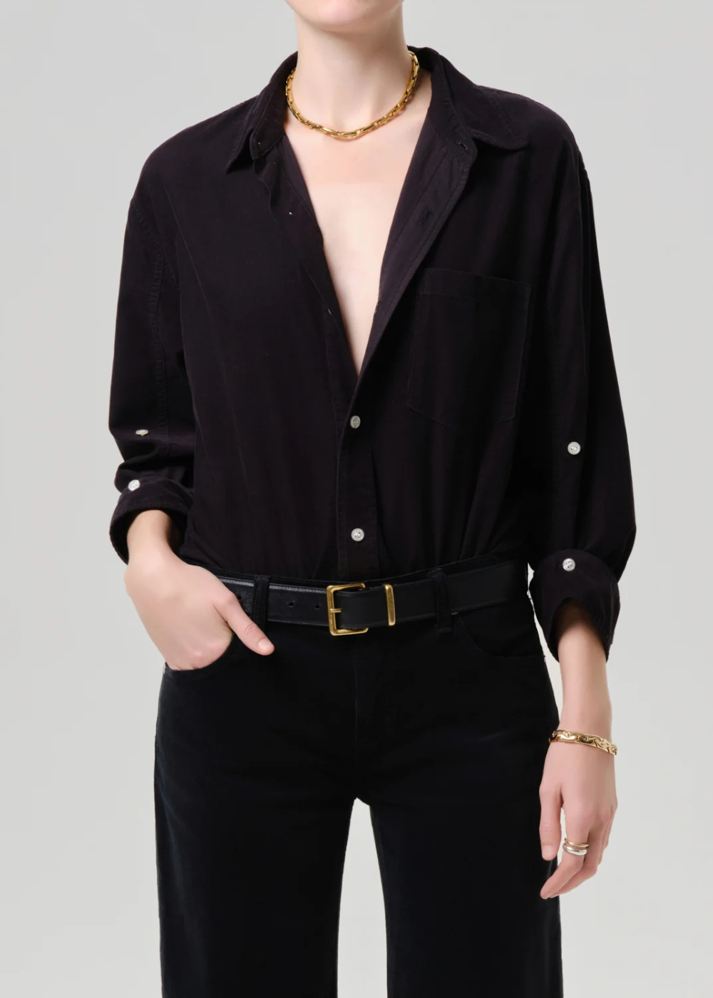 A model wearing a black long-sleeve button-up shirt with pearl-like button accents, paired with black trousers and a black belt with a gold buckle, against a light gray studio background.