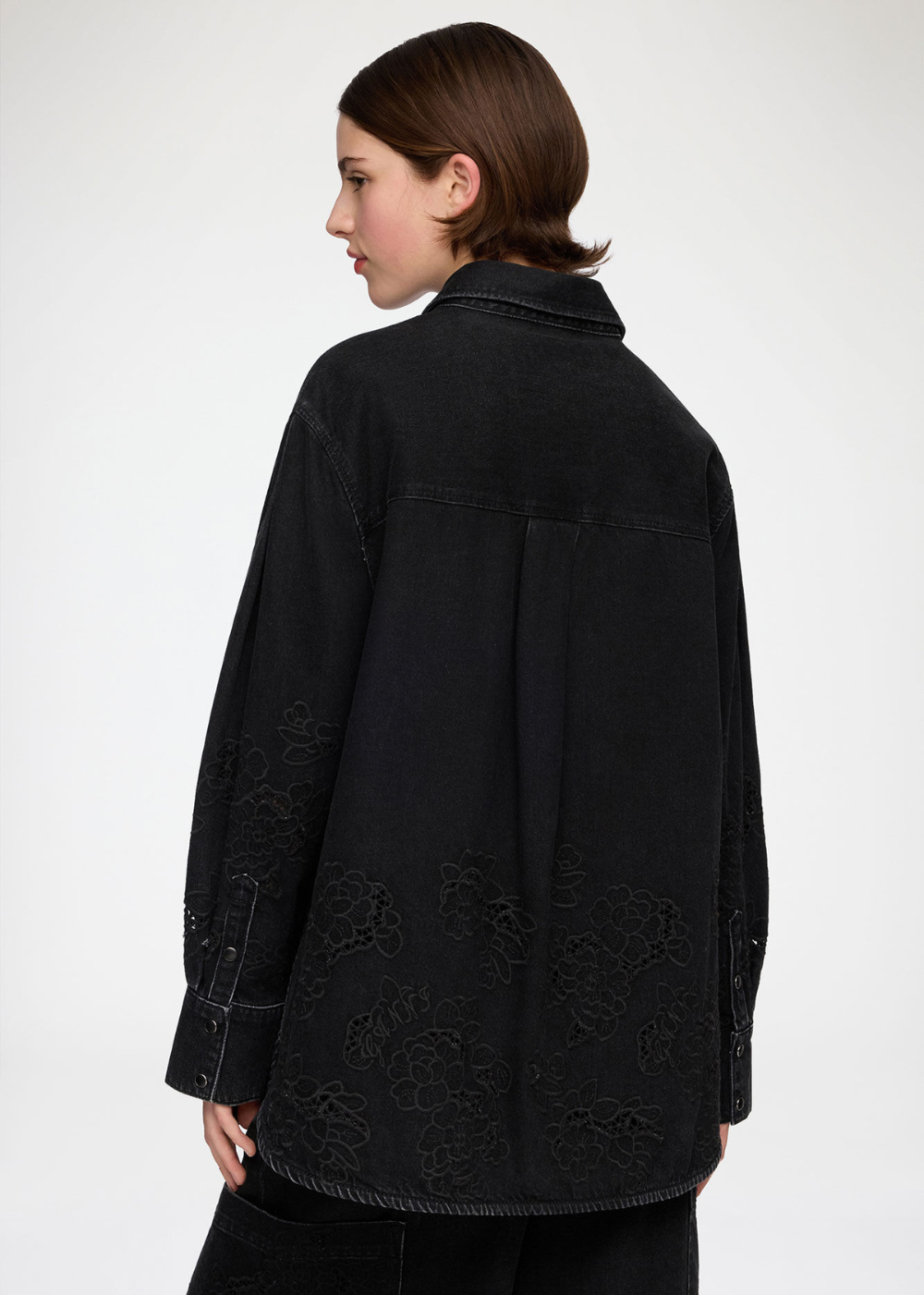 A model shown from the back wearing a black denim shirt jacket with tonal floral embroidery on the lower back and cuffs, set against a plain light gray studio background.