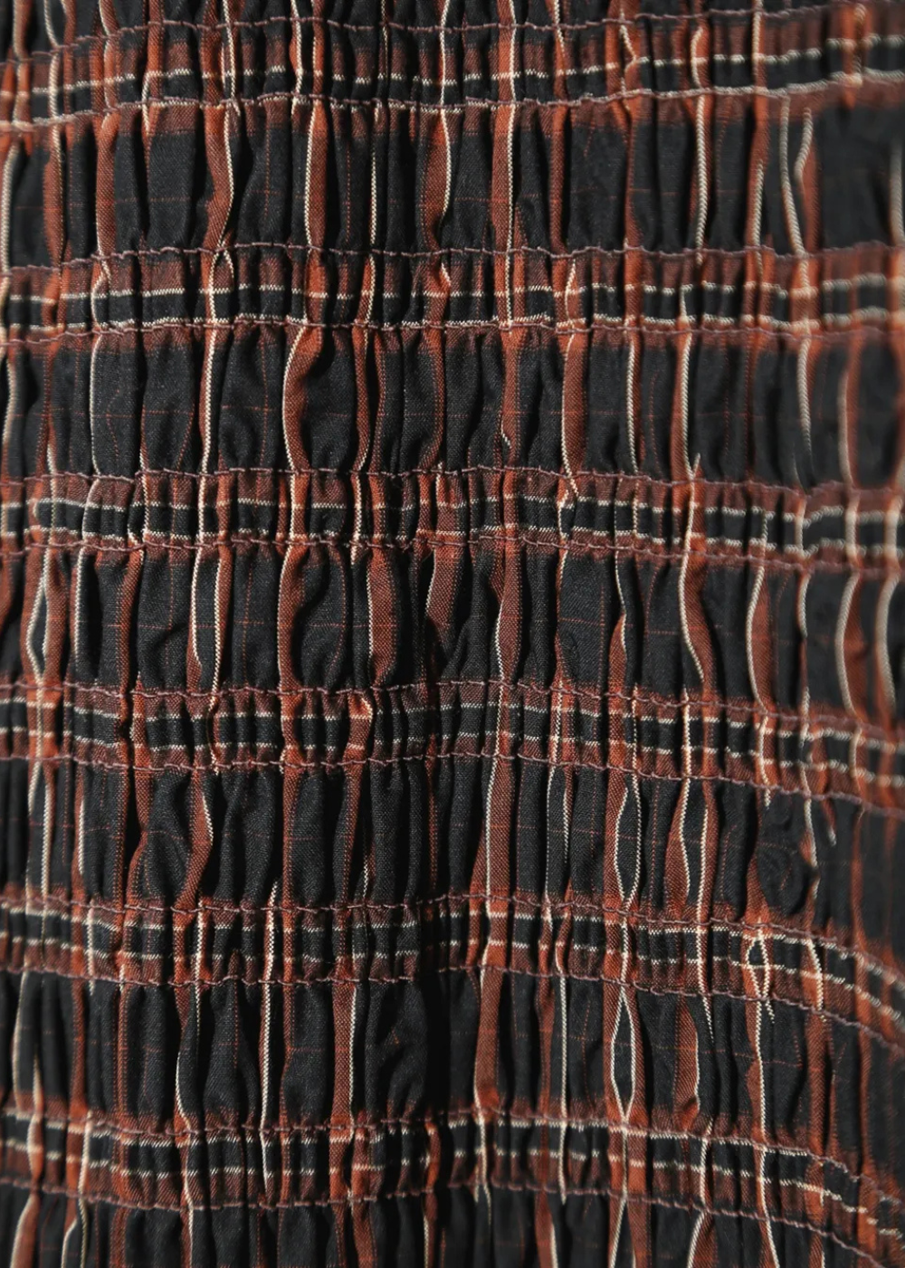 Close-up of a smocked fabric with a plaid pattern in black, rust orange, and thin white stripes.