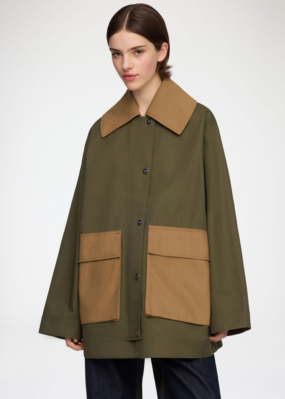 A model wearing an olive green utility jacket with contrasting tan collar, front buttons, and two large tan patch pockets, paired with dark blue jeans against a light gray studio background.