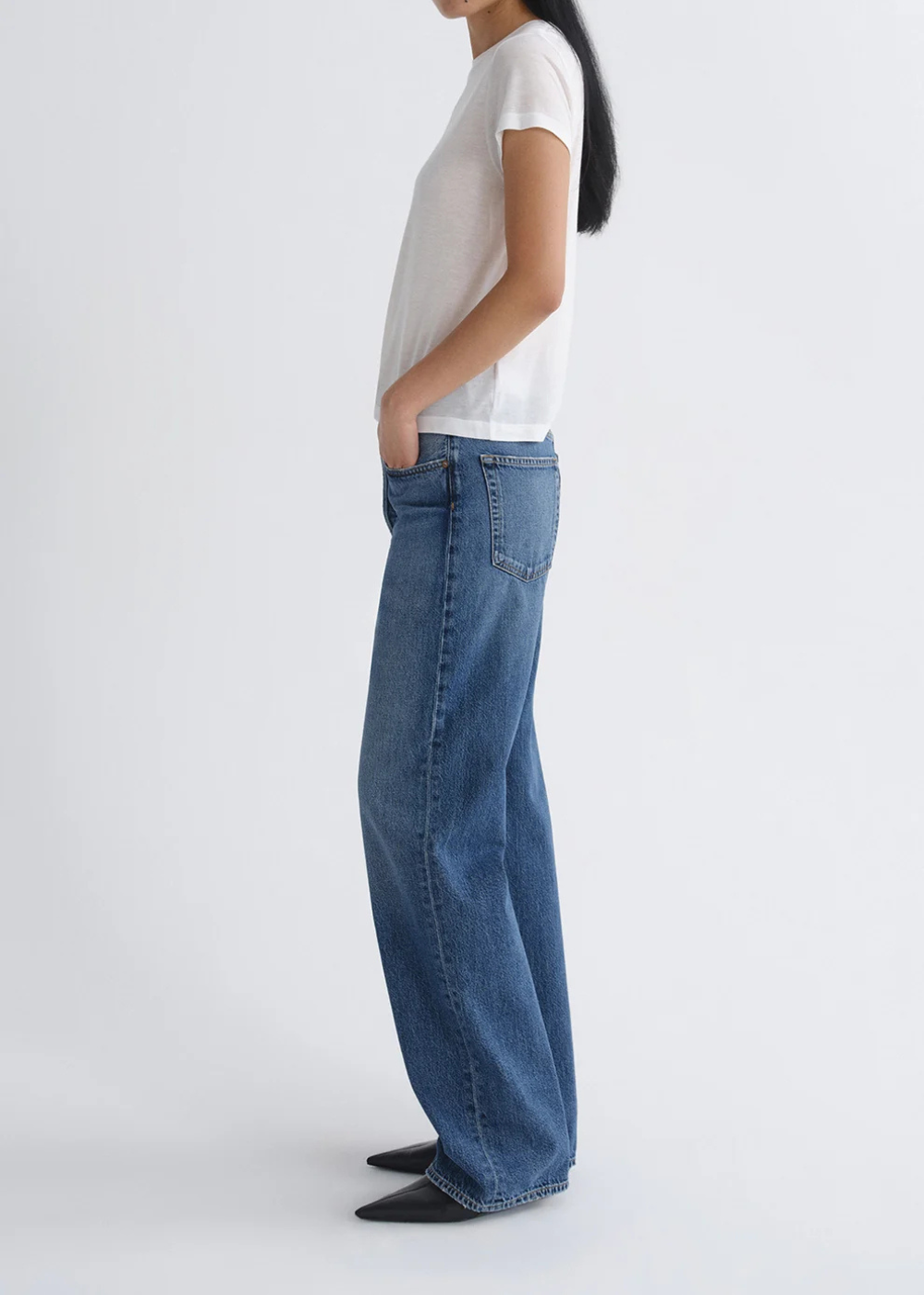 A side-profile view of a model wearing blue wide-leg jeans, a white short-sleeve t-shirt, and black pointed shoes against a plain light gray studio background.