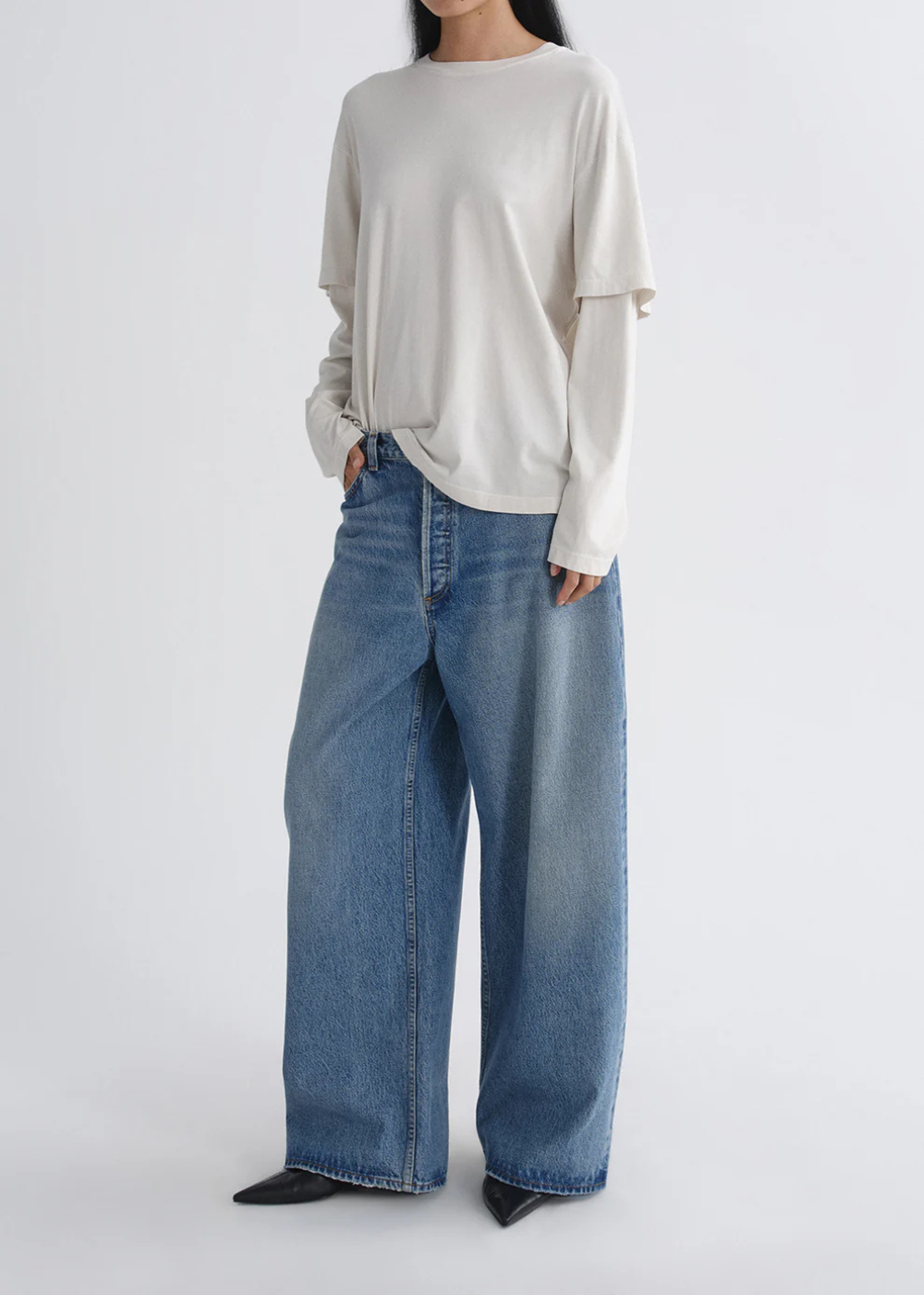 A model wearing a cream-colored long-sleeve top and wide-leg blue denim jeans, standing against a plain light gray studio background.