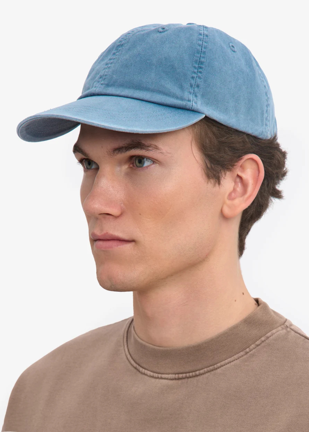 A man wearing a light blue washed baseball cap and a brown crewneck sweatshirt, shown in a side-profile portrait against a plain white background.