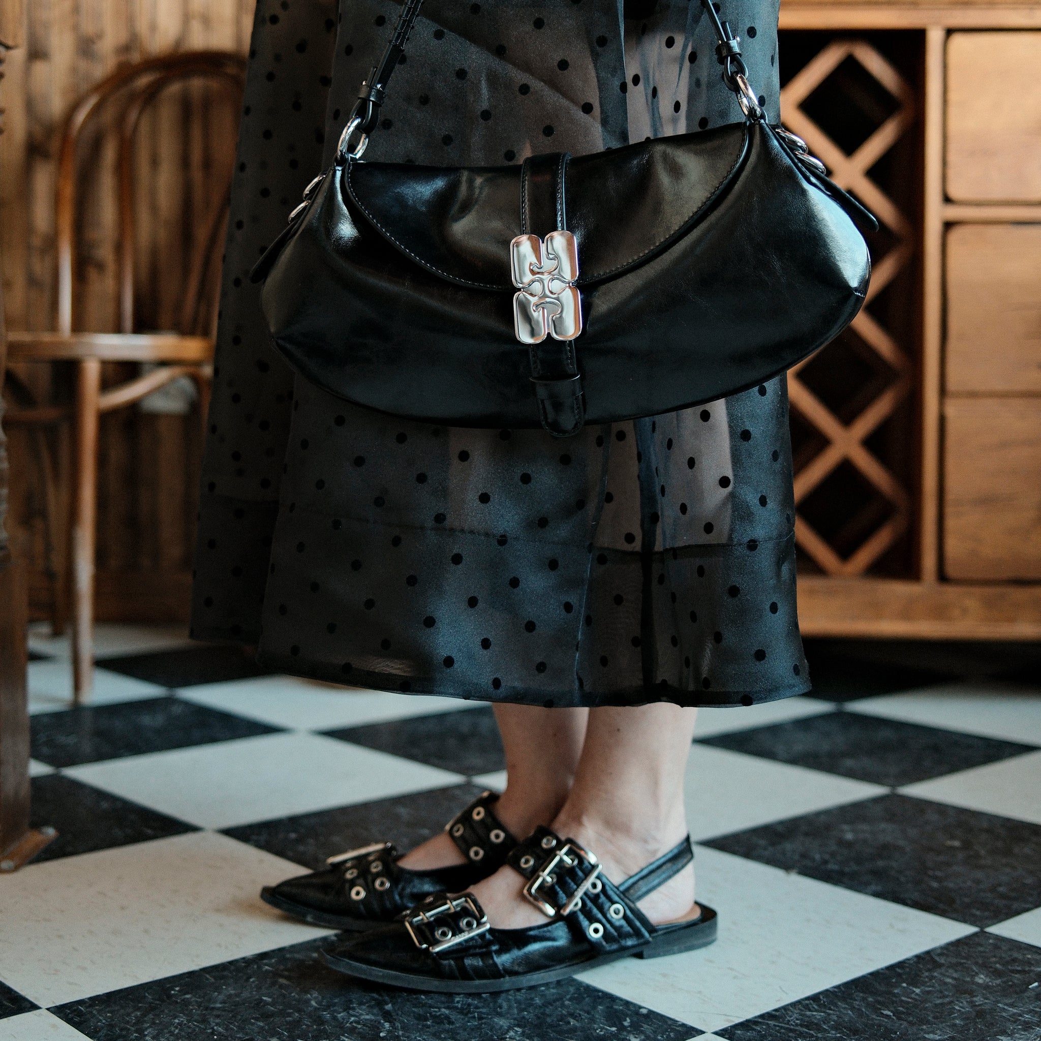 Person wearing a black polka dot dress and black sandals with silver buckles, holding a black handbag.