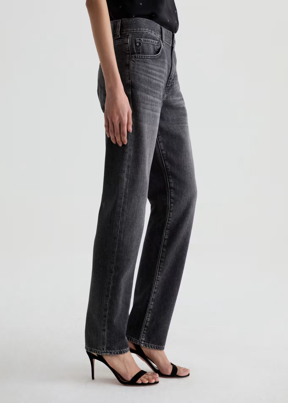 A model wearing a pair of dark gray washed straight-leg jeans with black strappy high-heeled sandals, shown from the waist down against a light gray studio background.