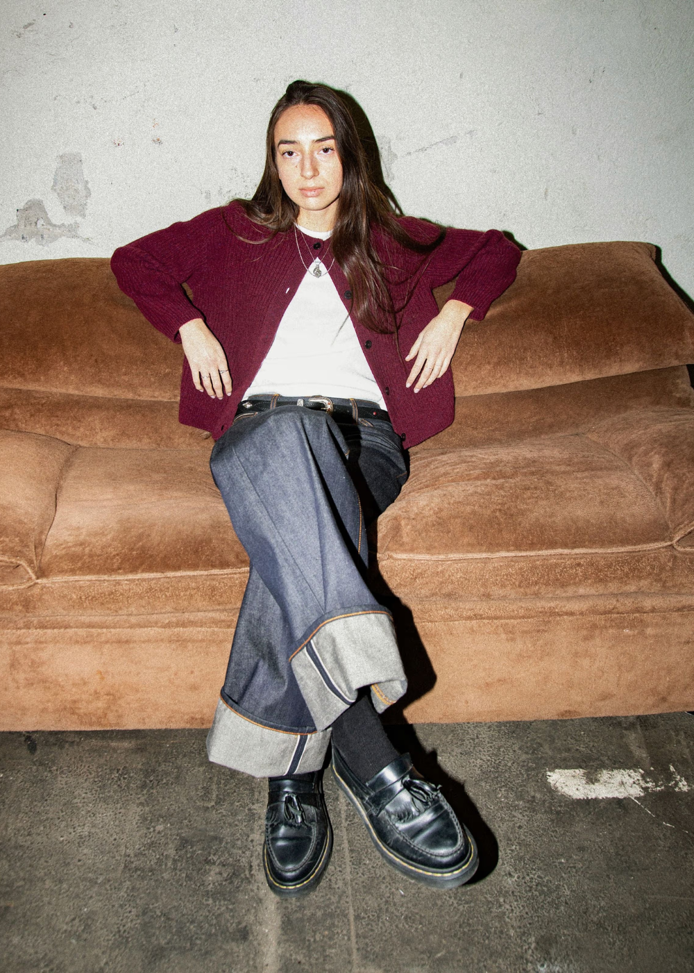 A model sits on a brown velvet sofa wearing a burgundy cardigan over a white t-shirt, dark wide-leg jeans with turned-up cuffs, black socks, and black leather loafers.