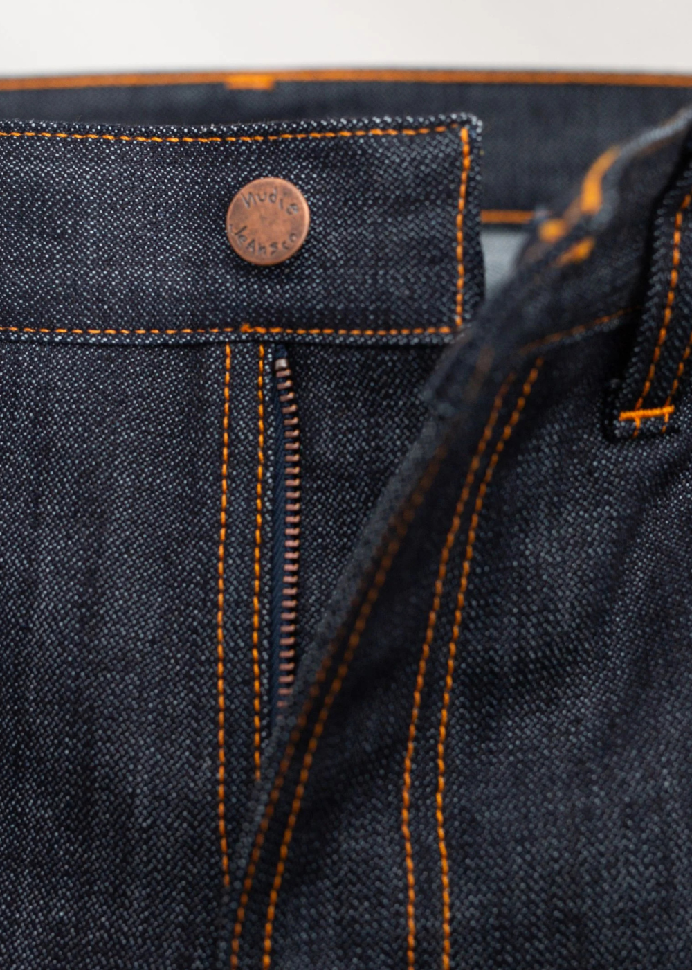 Close-up of dark indigo denim jeans showing the waistband, button closure, zipper, and orange contrast stitching.