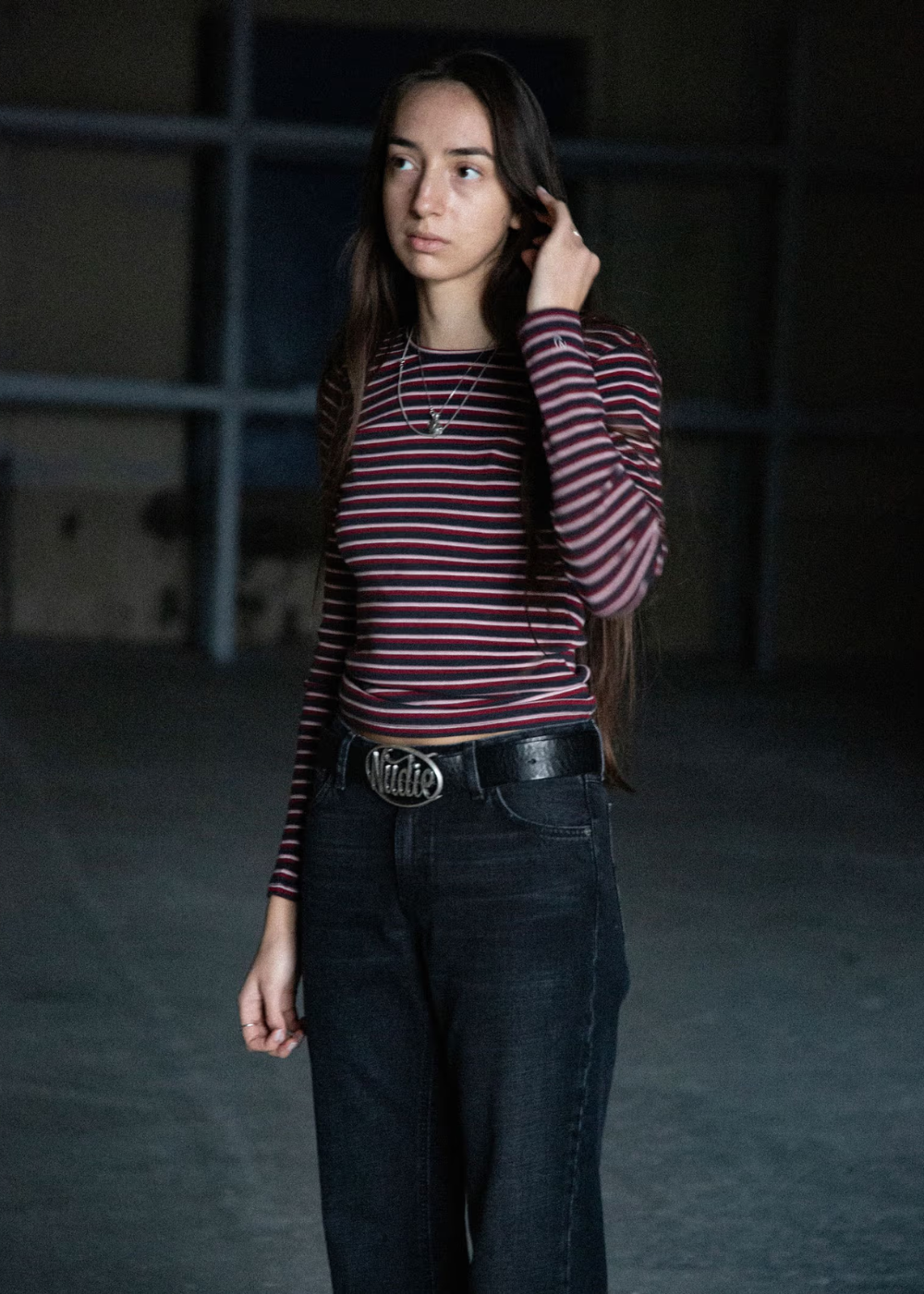 A woman wearing a burgundy and navy striped long-sleeve t-shirt, dark wash jeans, and a black belt with a silver buckle, standing in a dimly lit industrial setting.