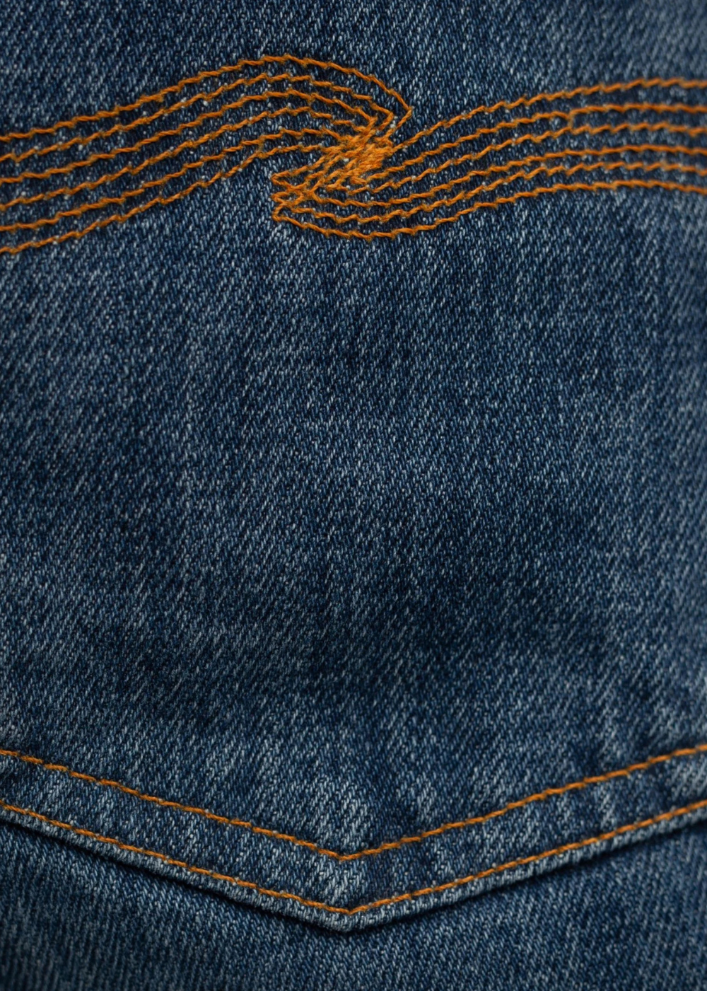 Close-up of a blue denim fabric showing the texture of the weave and orange contrast stitching along the pocket edge.