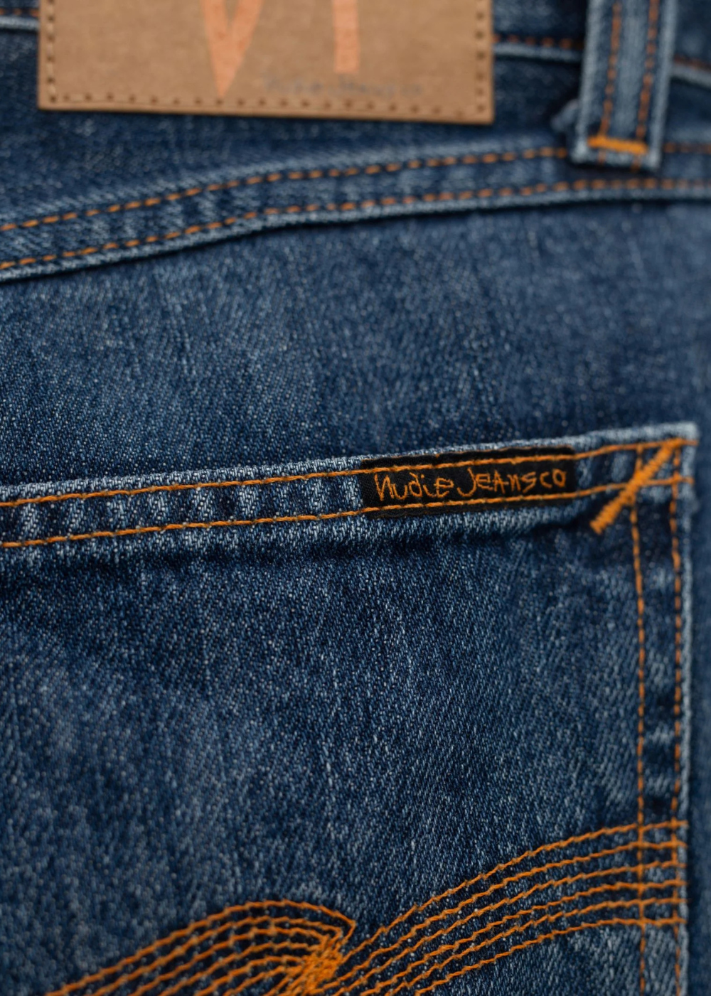 Close-up of blue denim jeans showing the back pocket with orange contrast stitching, a tan leather patch, and a visible 'Rude Jeansco' embroidered label.