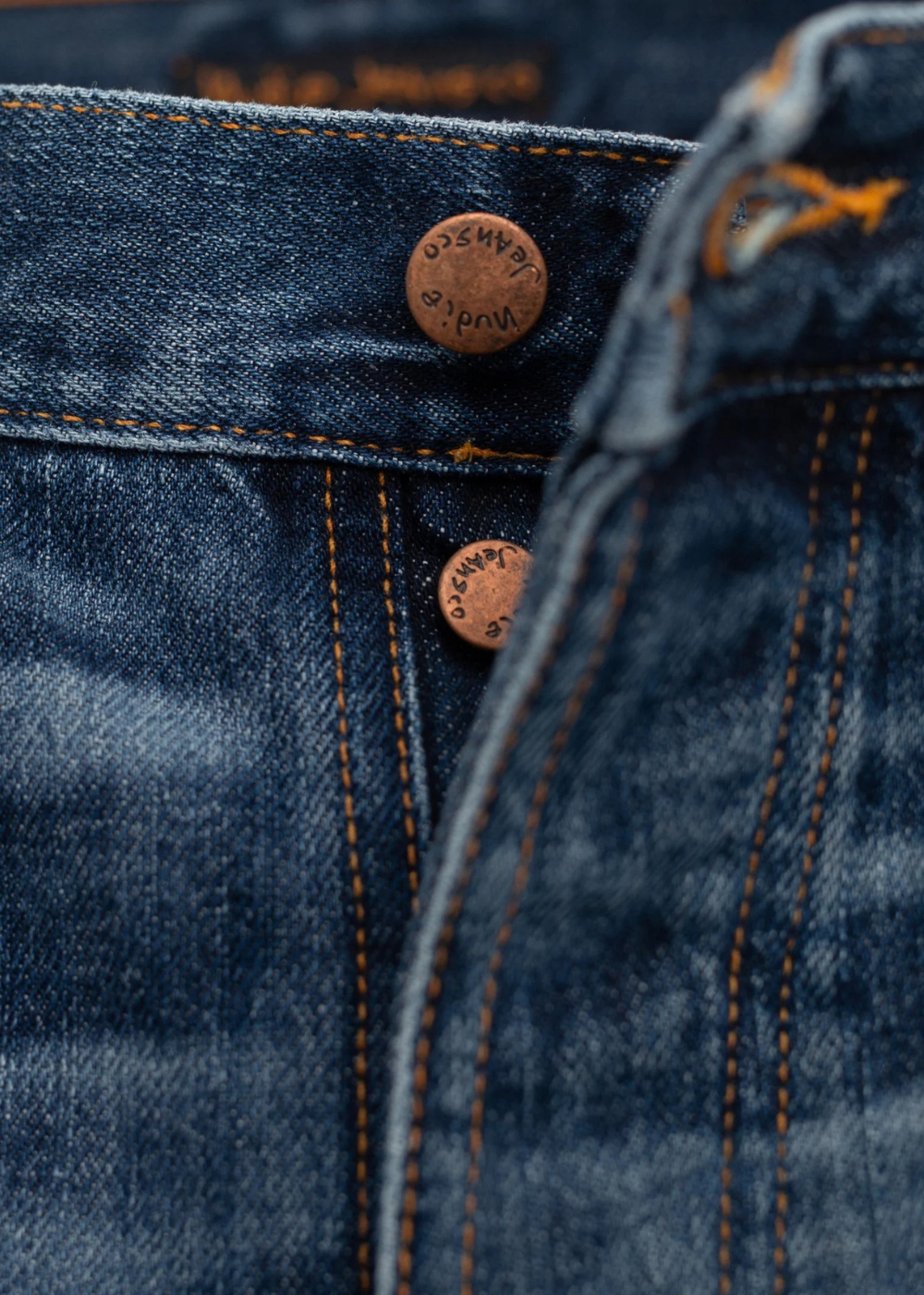 Close-up of dark blue denim jeans showing the button fly area with copper-toned metal buttons and orange contrast stitching.