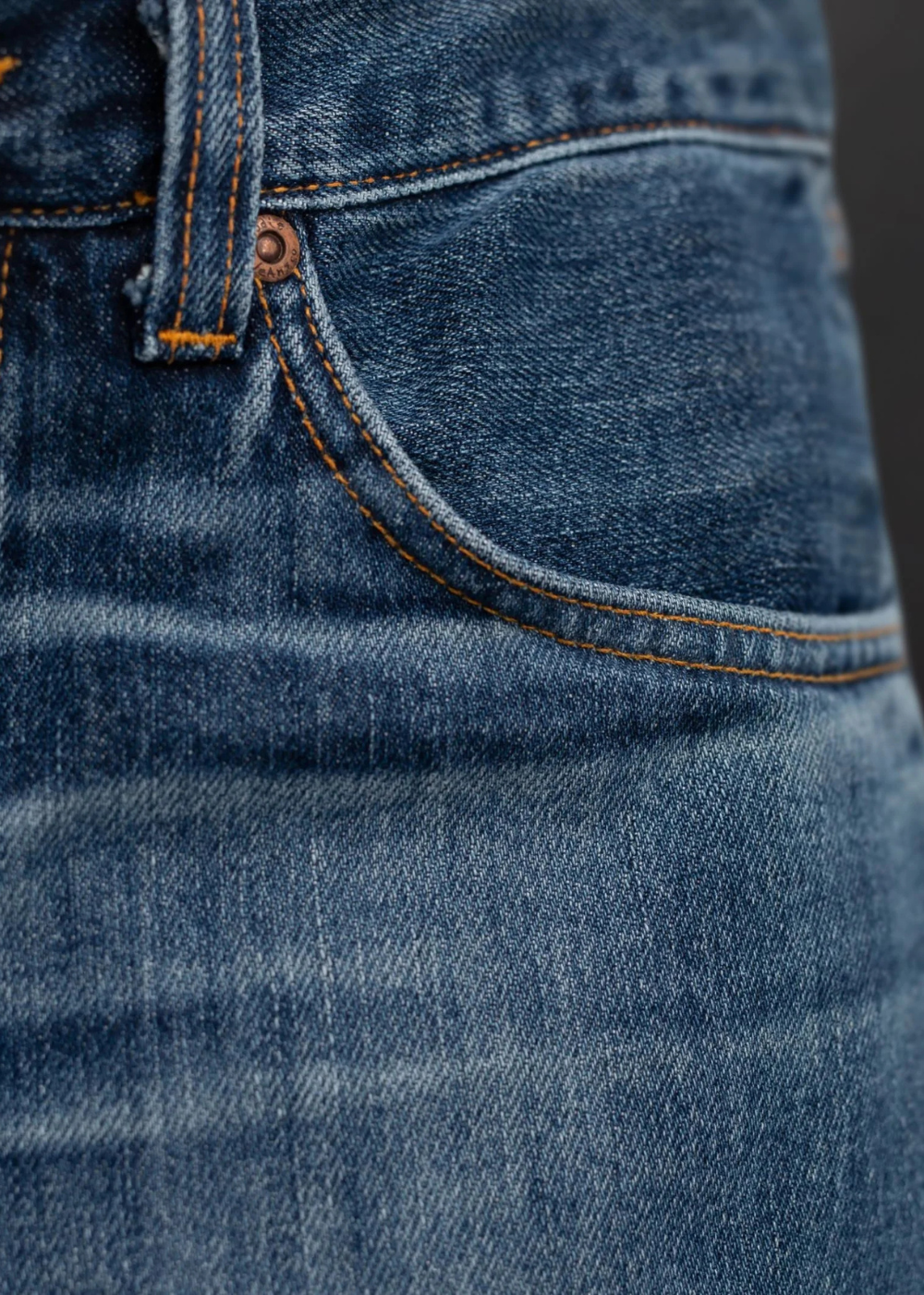 Close-up of a blue denim garment showing the front pocket, stitching, rivet, and waistband detail.