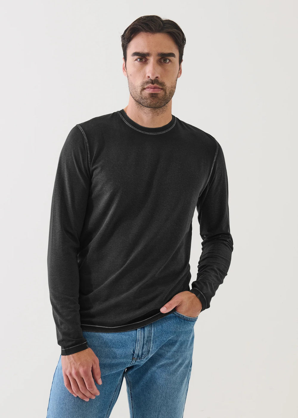 A man wearing a black long-sleeve crewneck shirt and blue jeans, standing against a plain light gray studio background.
