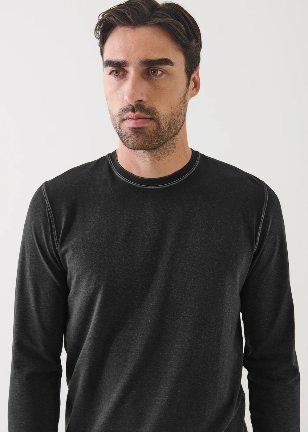 A man wearing a black long-sleeve crewneck shirt with subtle contrast stitching, photographed against a plain light gray background.