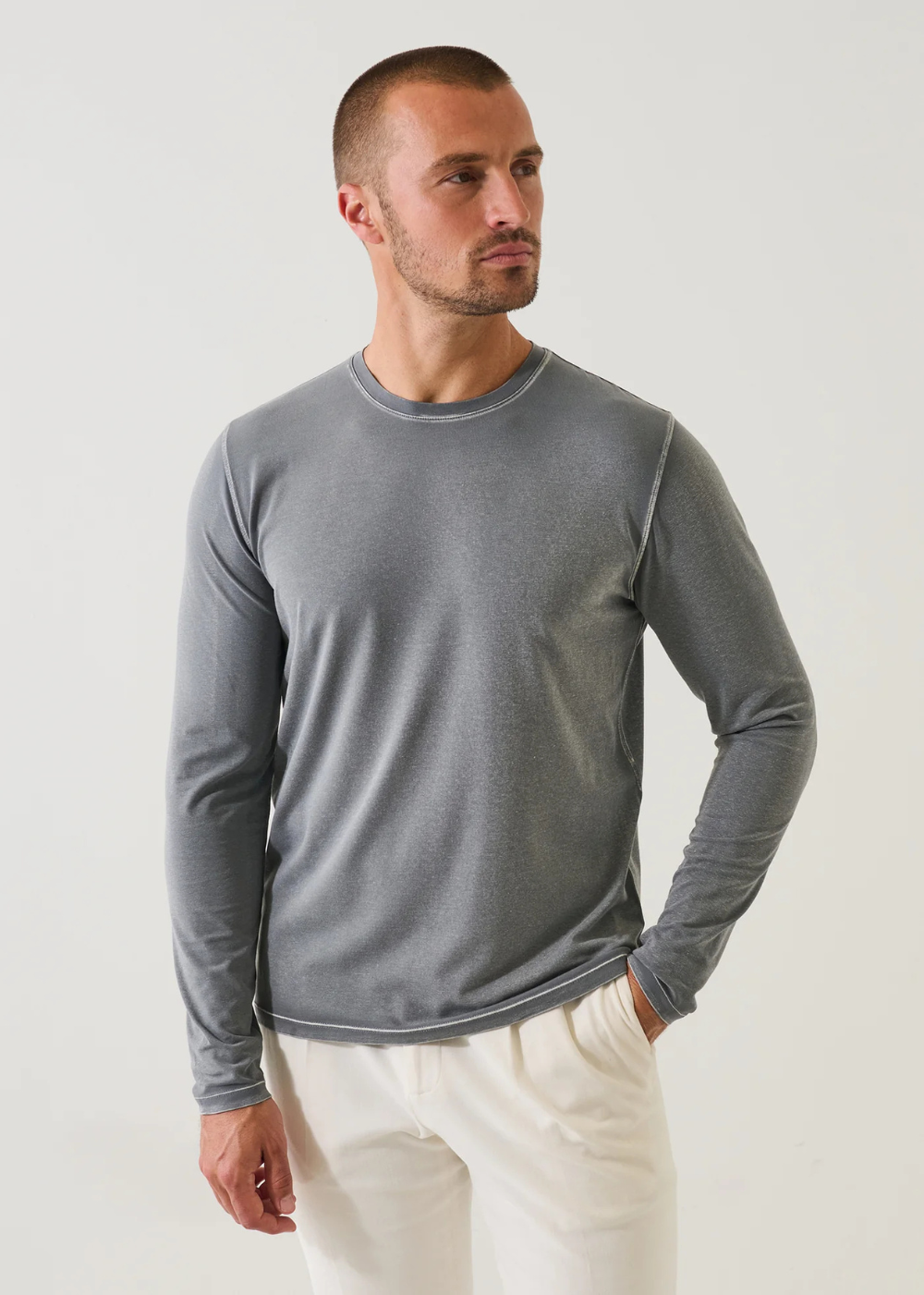 A man wearing a gray long-sleeve crewneck shirt and off-white pants, standing against a light gray studio background.