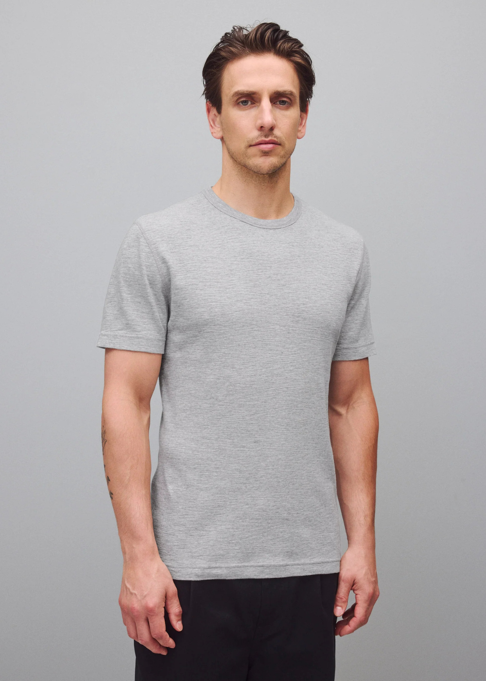 A man wearing a light gray short-sleeve crewneck t-shirt and black pants, standing against a solid gray studio background.