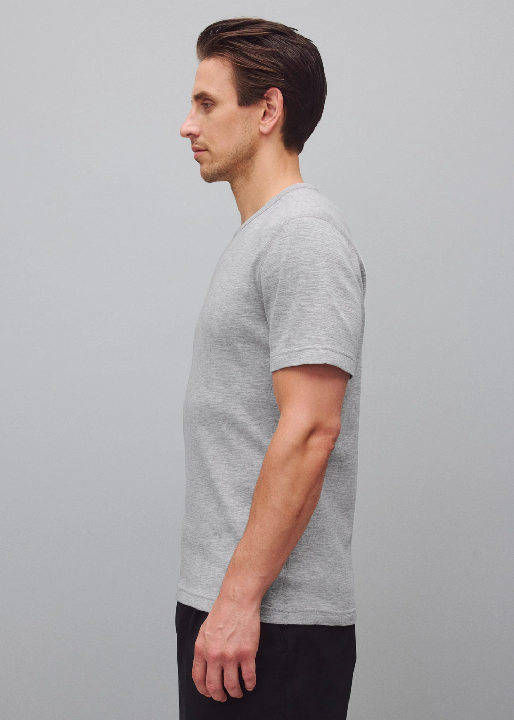 A man in side profile wearing a light gray short-sleeve crewneck t-shirt and dark pants, standing against a solid gray studio background.