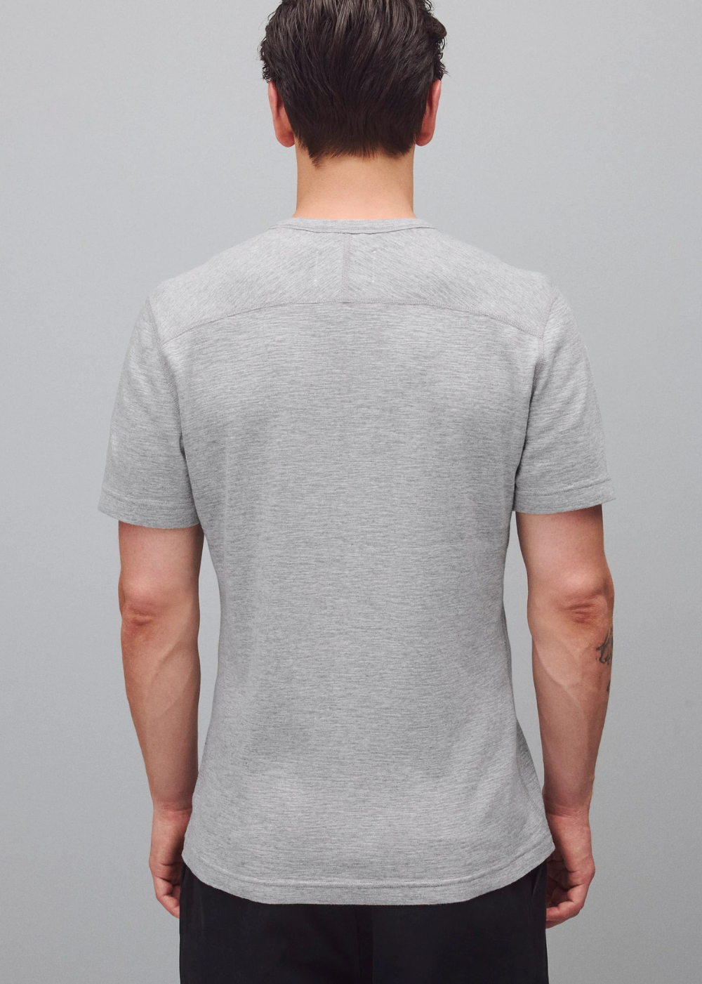 A man viewed from the back wearing a light gray heather crewneck t-shirt and black pants, standing against a solid gray studio background.