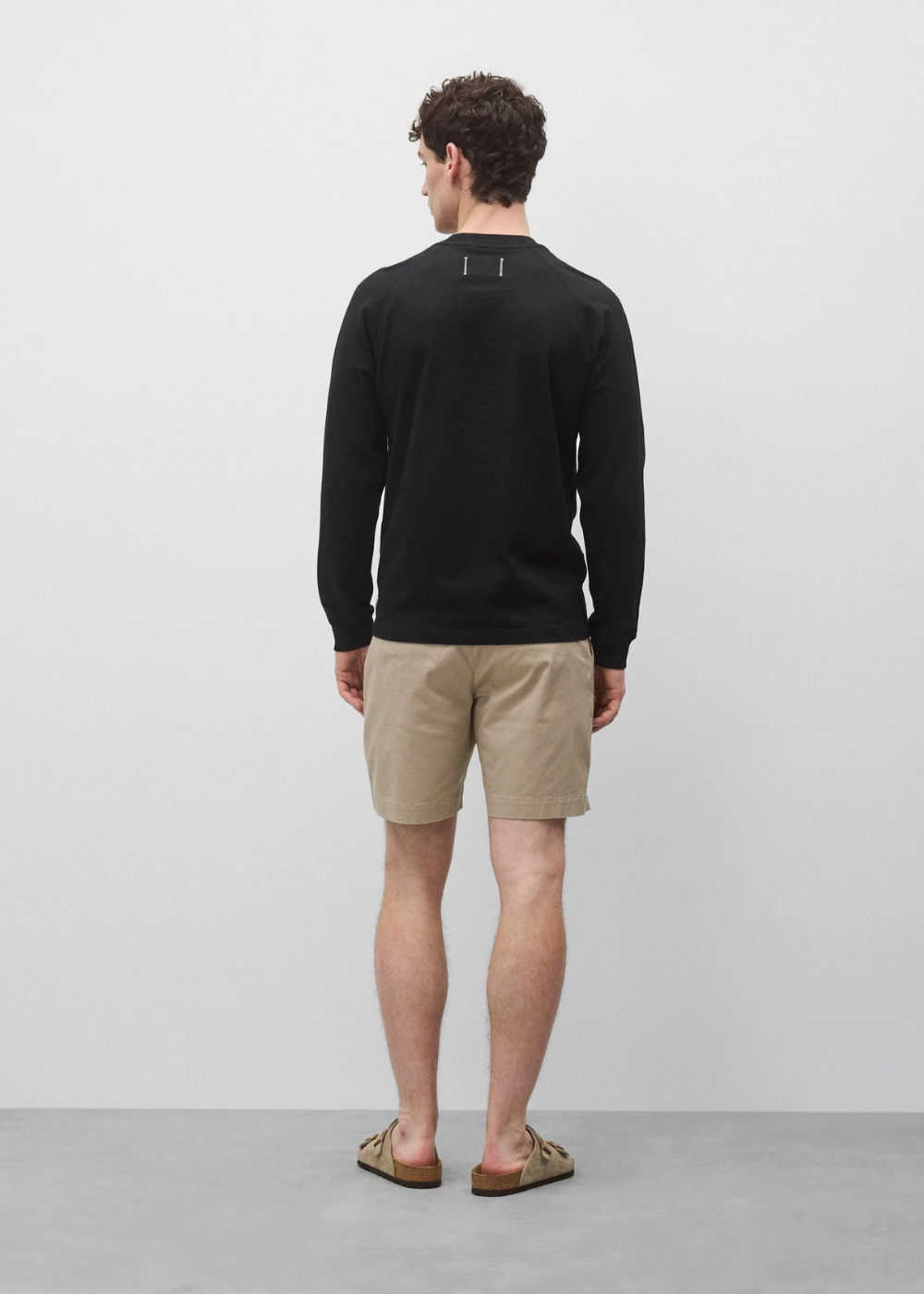 A man standing with his back to the camera, wearing a black long-sleeve crewneck sweater, beige shorts, and brown sandals, photographed in a minimalist studio setting.