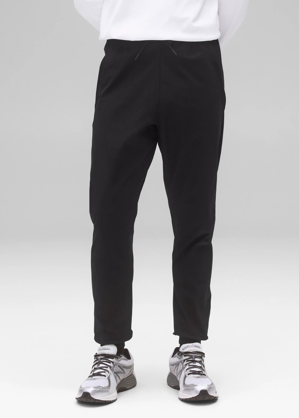 Black slim-fit jogger pants shown on a model from the waist down, paired with a white sweatshirt and gray running shoes against a light gray studio background.