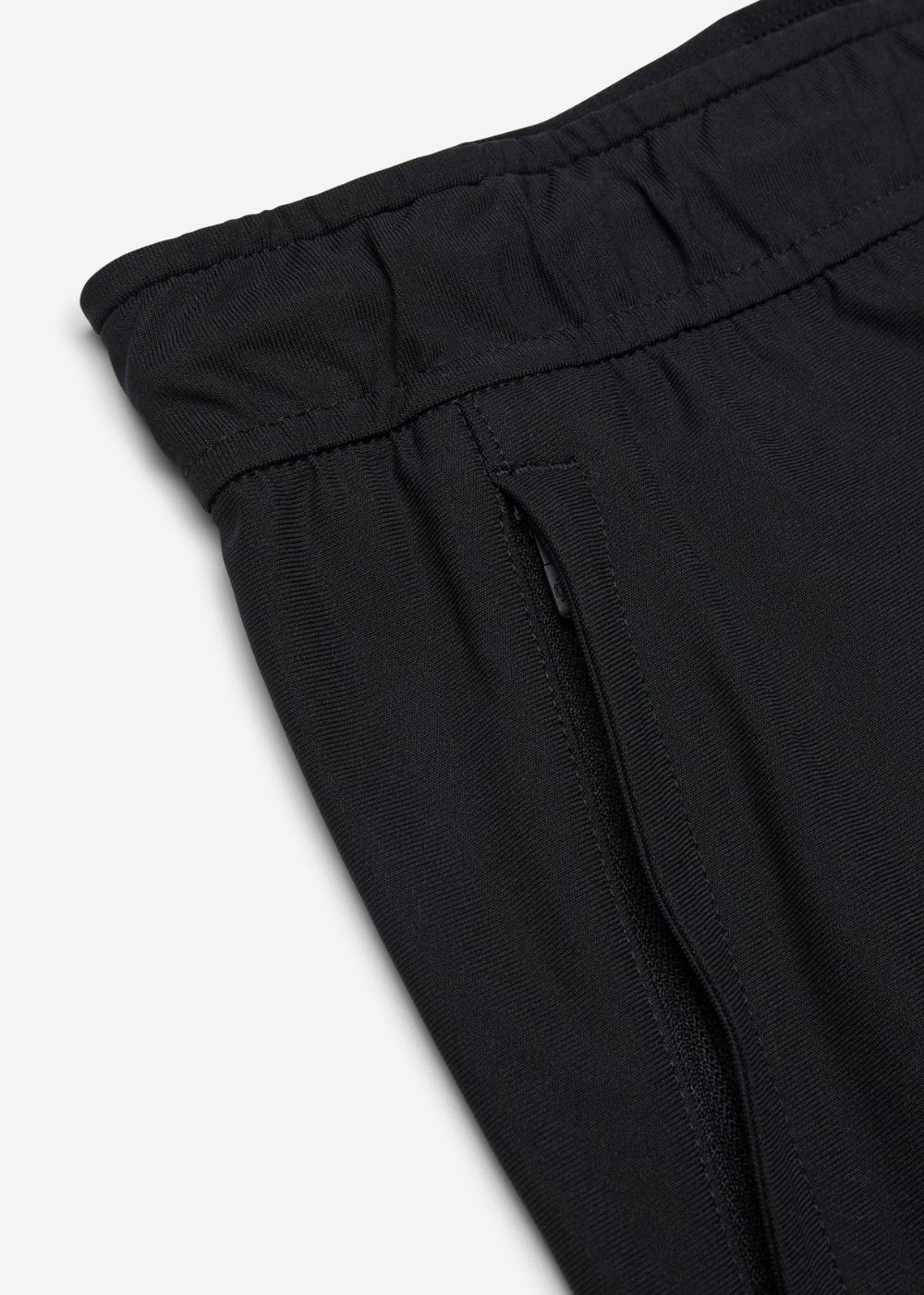 Close-up detail of black trousers showing the elastic waistband, stitched seam, and side pocket on a light gray background.